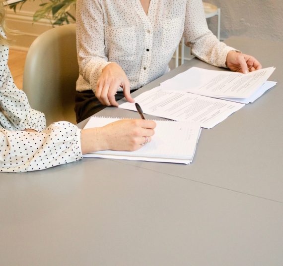 Cliente firmando unos papeles mientras está siendo asesorada por su abogada de confianza. Ambas llevan camisas de color blanco con topos negros.